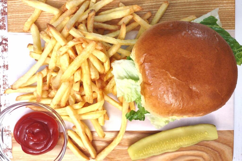 top down view of hamburger, fries, ketchup, and a pickle on a wooden cutting board