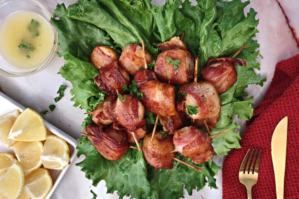 overhead view of bacon wrapped scallops on lettuce with lemon wedges and melted butter to the side