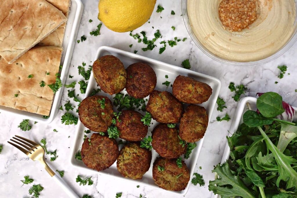 overhead view of 11 falafel on a square plate with pita, hummus, and lemon on the side