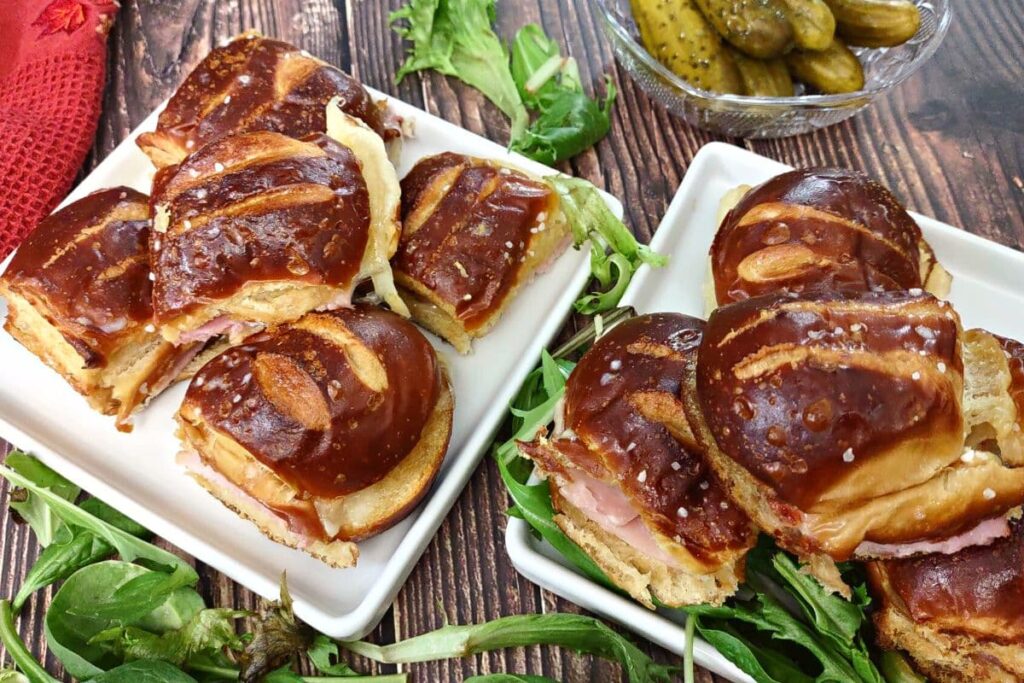 two plates of ham and cheese sliders on hawaiian pretzel rolls with pickles and greens