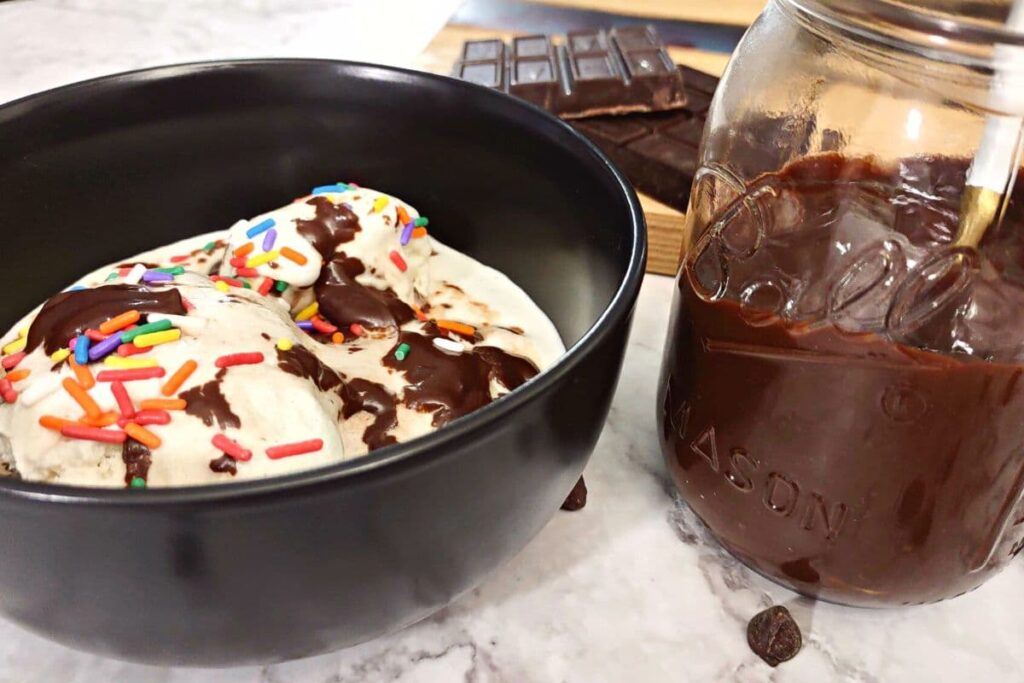 homemade chocolate sauce in a jar next to a bowl of vanilla ice cream with sprinkles