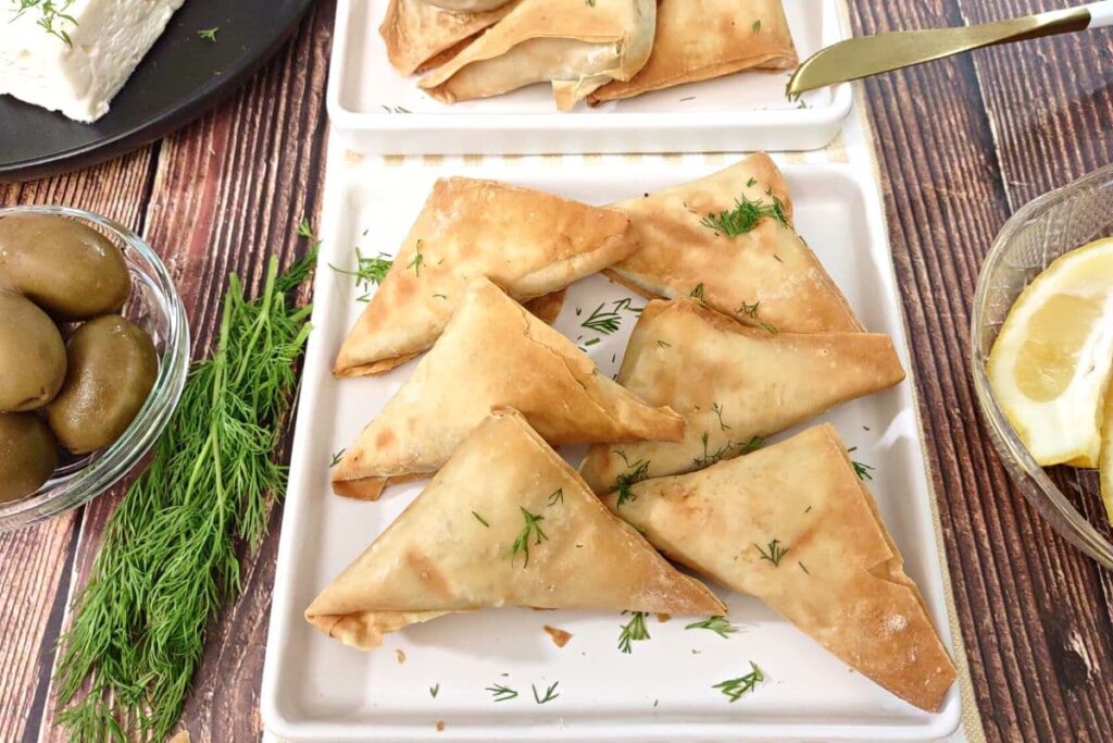 flaky air fried spanakopita on a white square plate with lemons and olives on the side