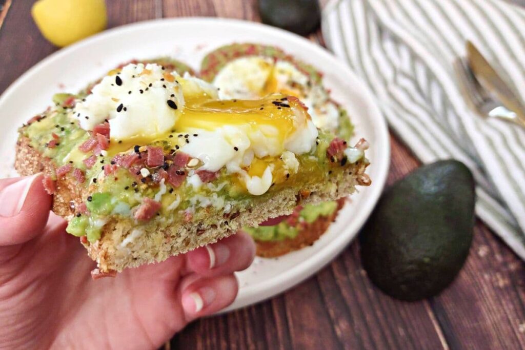 holding up a slice of avocado toast with a cracked runny egg
