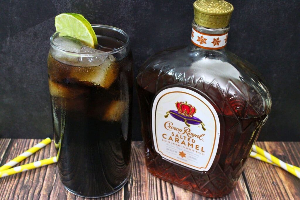 highball glass of crown salted caramel and coke with a lime slice next to the whiskey bottle