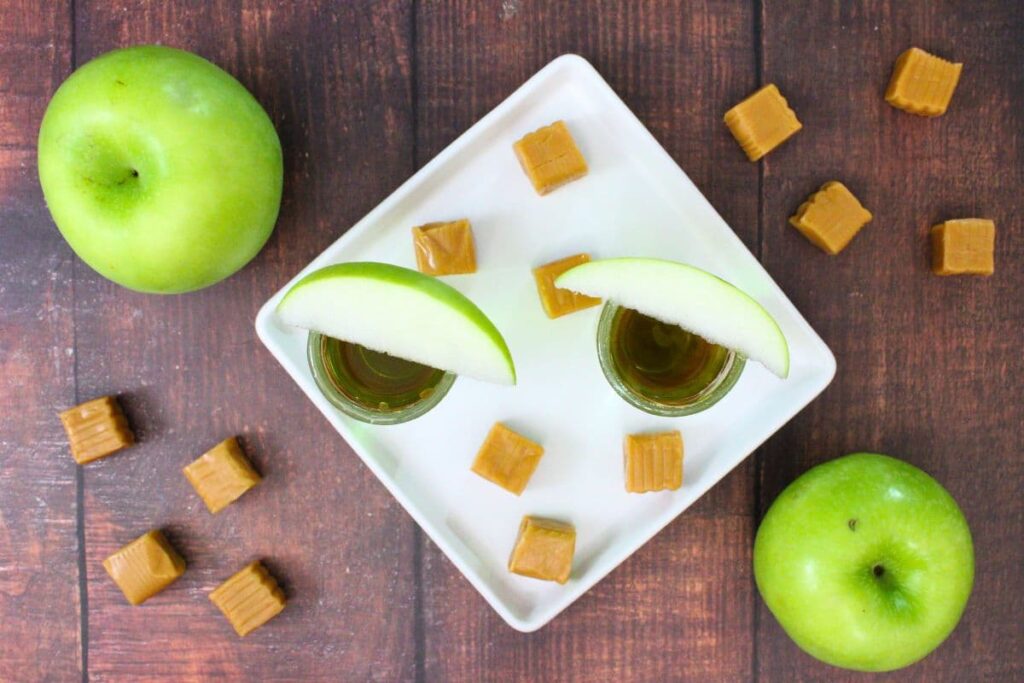 overhead view of two caramel apple shots with crown next to caramel squares and fresh apples