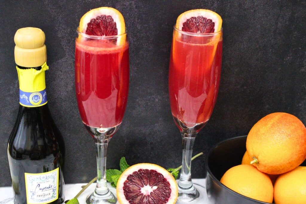 blood orange mimosas in champagne flutes with bottle and fresh oranges next to it