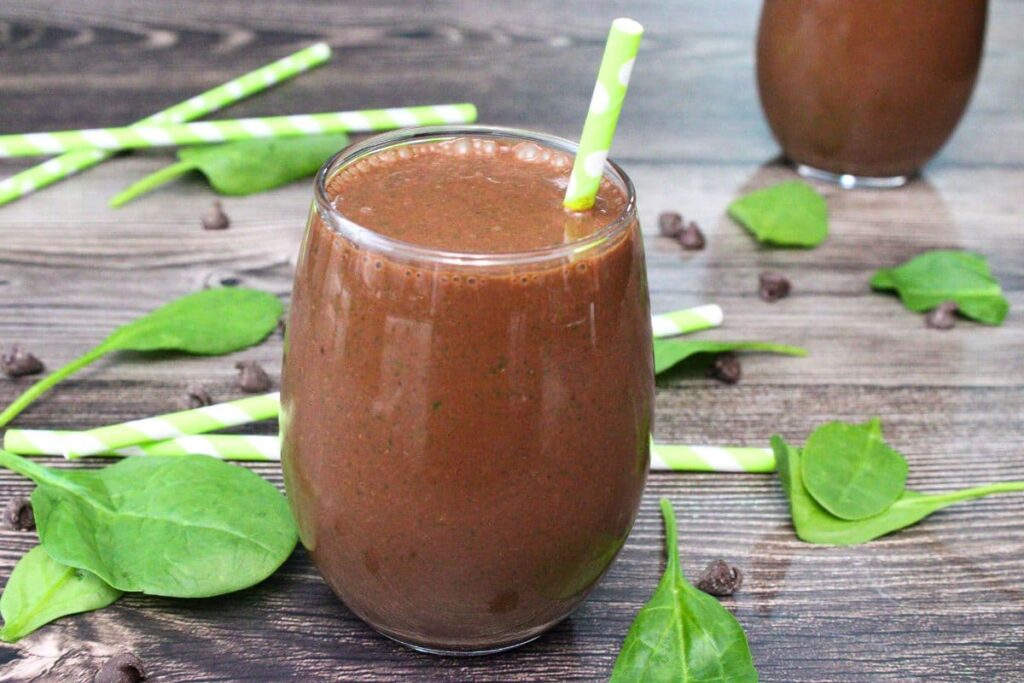 chocolate peanut butter spinach smoothie with a paper straw surrounded by spinach leaves