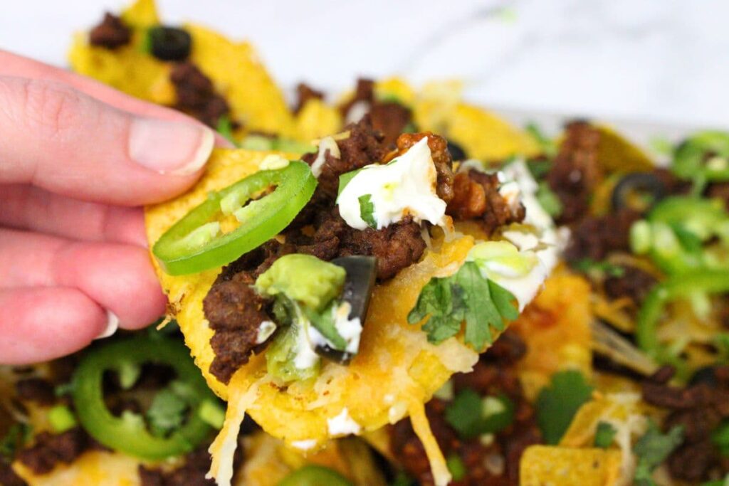 holding up an air fryer nacho chip with ground beef, jalapeno, sour cream, and black olives