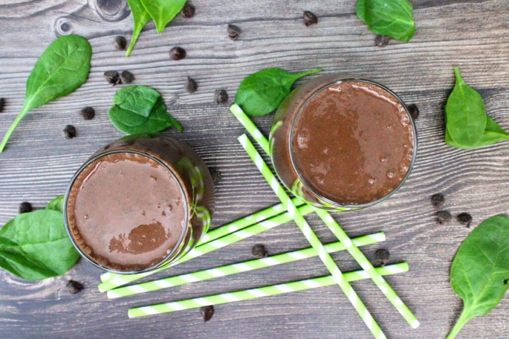 overhead view of two glasses of chocolate peanut butter spinach smoothie with straws and spinach surrounding