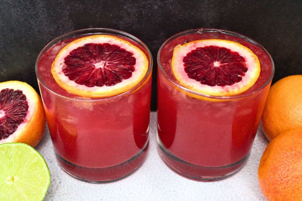 two glasses filled with blood orange paloma in front of a dark background