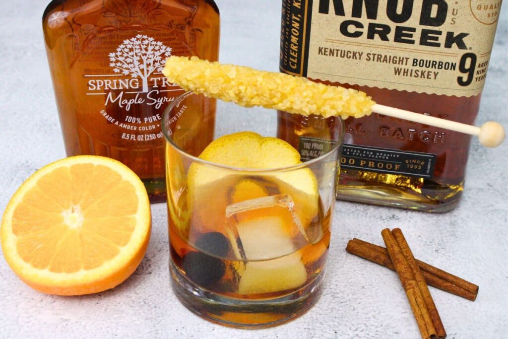 maple old fashioned with a rock candy stick on top and orange and cinnamon on the sides