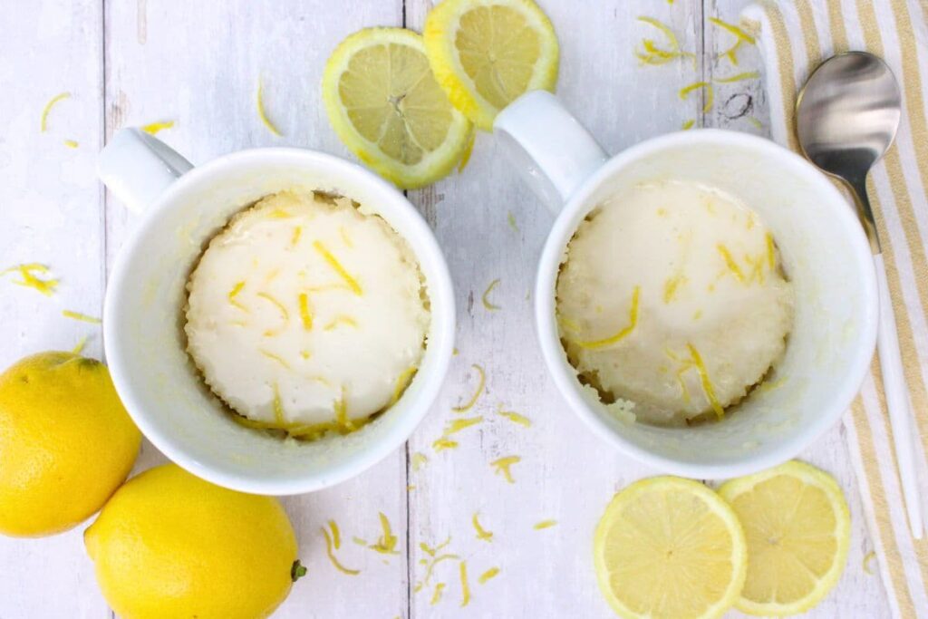 overhead view of two lemon mug cakes on white wood with fresh lemons surrounding