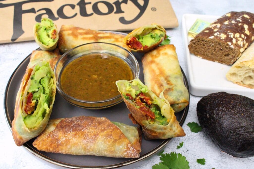 cheesecake factory copycat avocado egg rolls surrounding a plate with dipping sauce and a fresh avocado
