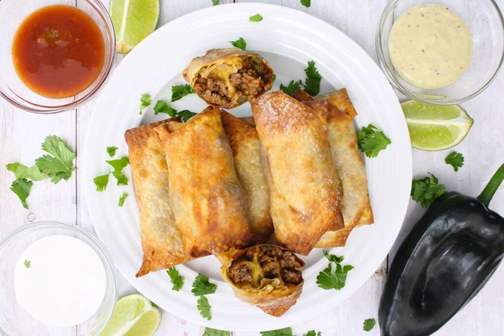 overhead view of air fryer taco egg rolls stacked on a plate with dipping sauces on the side