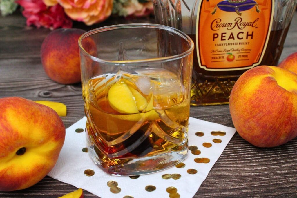 crown royal peach old fashioned drink in front of the bottle next to whole peaches