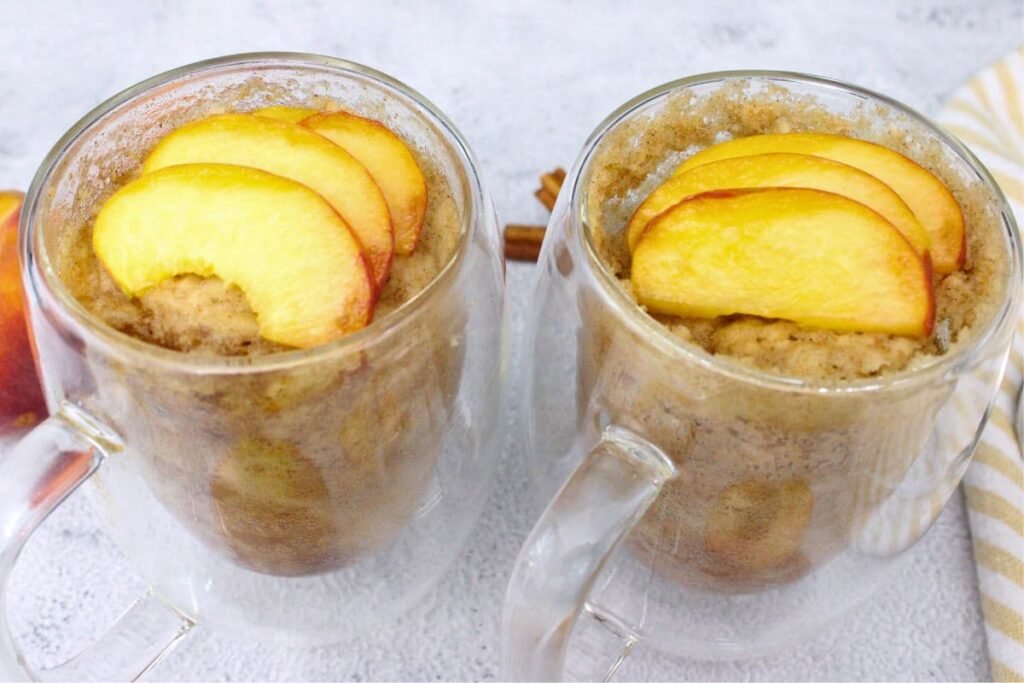 two peach mug cakes in see through mugs with fresh peach slices on top