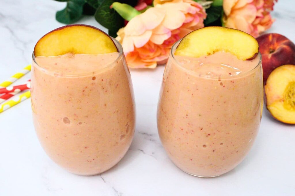 two peach strawberry mango smoothies next to each other with a peach wedge on top