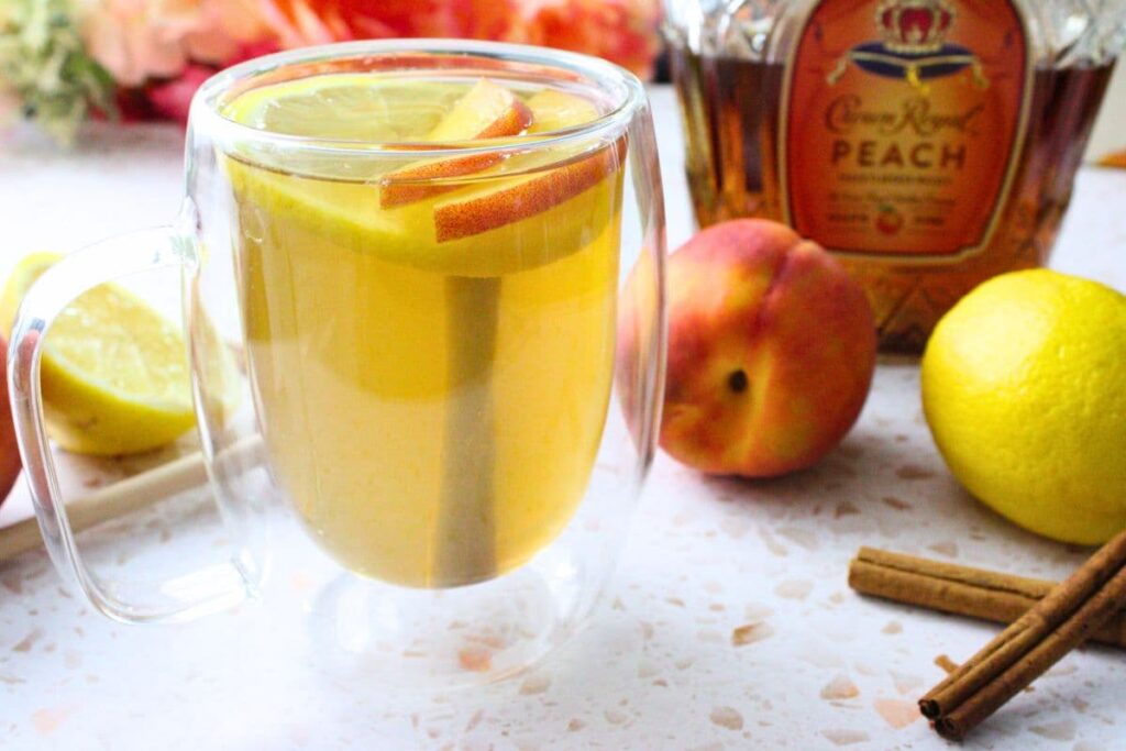 crown peach hot toddy drink surrounded by fresh fruit and cinnamon sticks