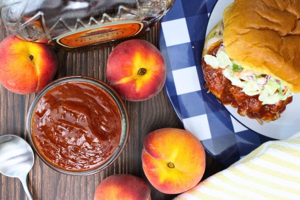 overhead view of a jar filled with whiskey peach bbq sauce next to a pulled pork sandwich