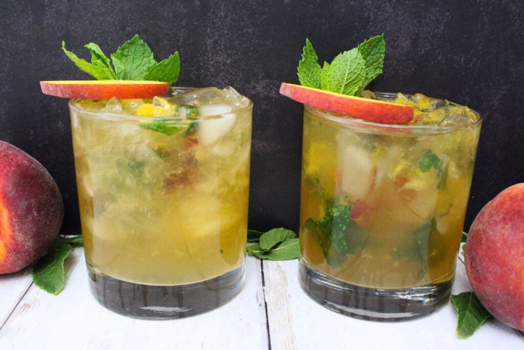 two peach mint julep cocktails on a white wooden table against a dark background