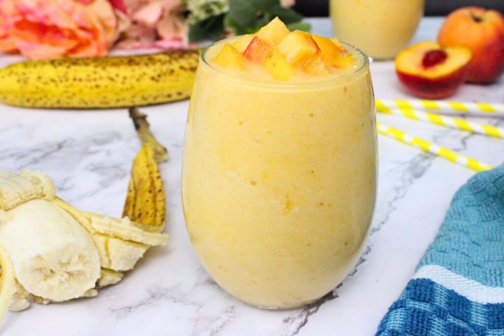 peach banana smoothie with peach chunks on top next to half a fresh banana