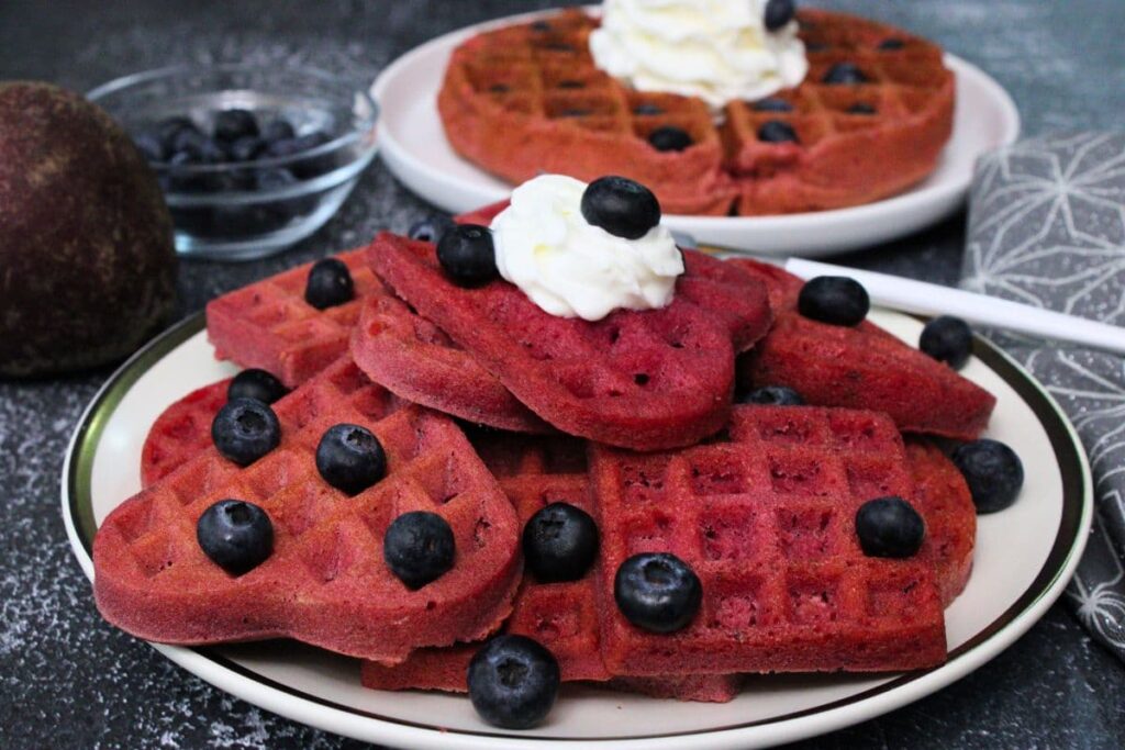 mini beetroot waffles stacked on a plate with whipped cream and a belgian waffle behind