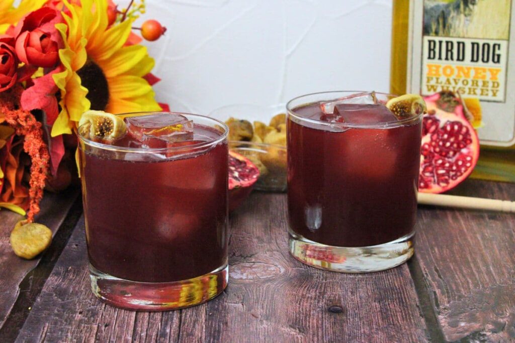 two gladiator cocktails in front of honey whiskey bottle and fresh pomegranates