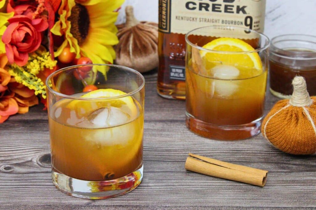 A close-up of a cocktail in a clear glass garnished with a round orange slice and a cinnamon stick. In the background, there is a knitted pumpkin decoration on a wooden surface. Another identical glass is partially visible.