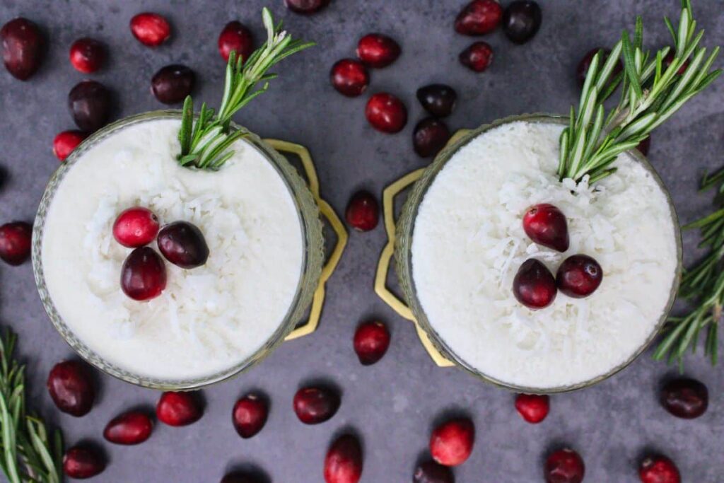 overhead view of two white christmas punch cocktails topped with cranberry, coconut, and rosemary
