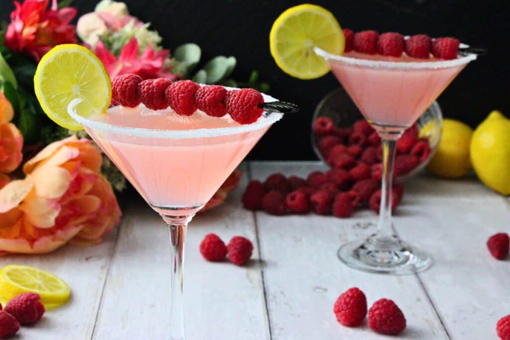 two pink lemon drop martinis garnished with fresh lemon and raspberries on white wooden table