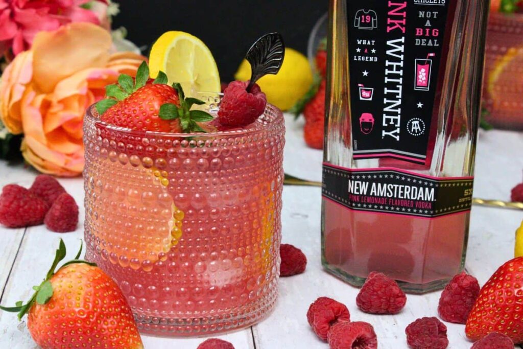 glass of pink whitney jungle juice next to vodka bottle surrounded by fresh fruit