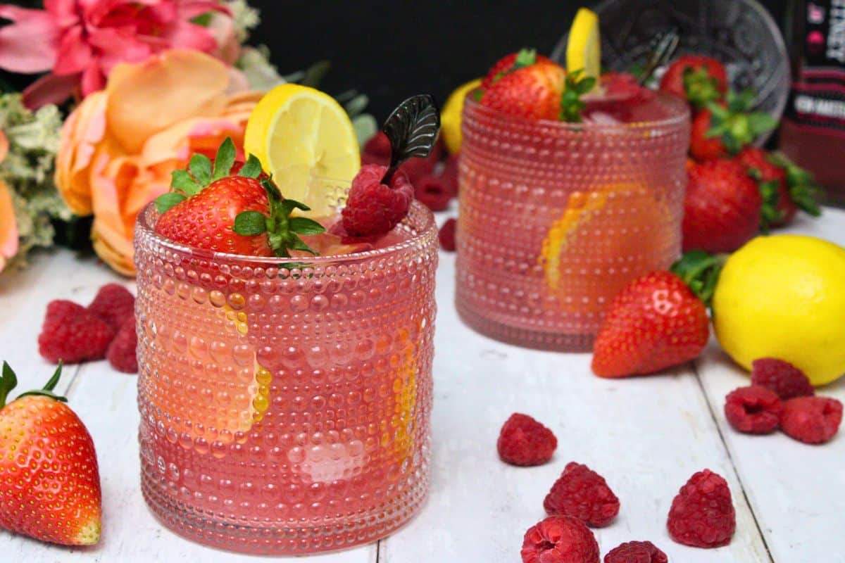 two glasses of pink whitney jungle juice on a white wooden table surrounded by fresh fruit and flowers