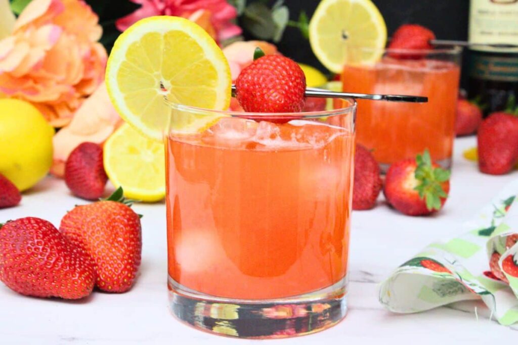 closeup of strawberry whiskey sour garnished with lemon slice and strawberry surrounded by fresh fruit with another drink in the background
