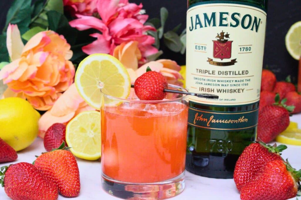 strawberry whiskey sour cocktail surrounded by fresh strawberries and lemon next to whiskey bottle