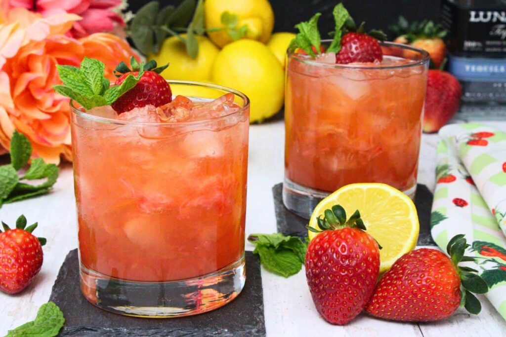 two strawberry tequila smash drinks on slate coasters surrounded by fresh strawberries and lemon