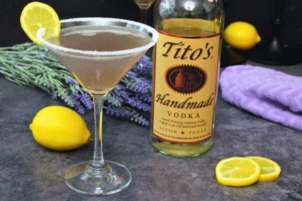 lavender lemon drop cocktail close to a bottle of vodka on a dark counter surrounded by lavender and lemons