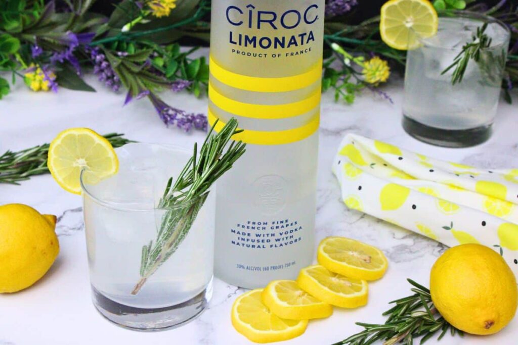 sparkling lemonade cocktail with a rosemary sprig next to tall vodka bottle surrounded by fresh lemons