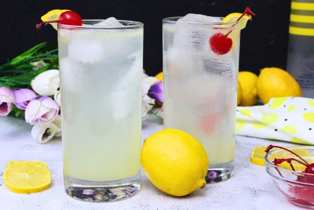 two tall lemon vodka collins drinks garnished with lemon and cherry surrounded by fresh lemons