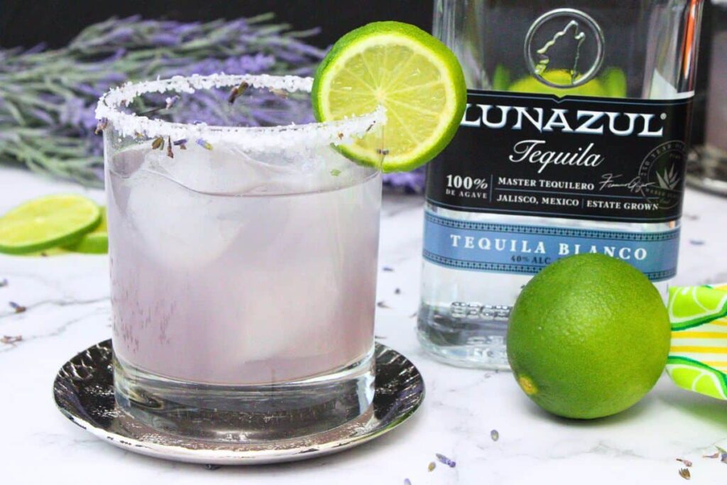 lavender margarita with lime slice next to tequila bottle surrounded by fresh lavender and lime