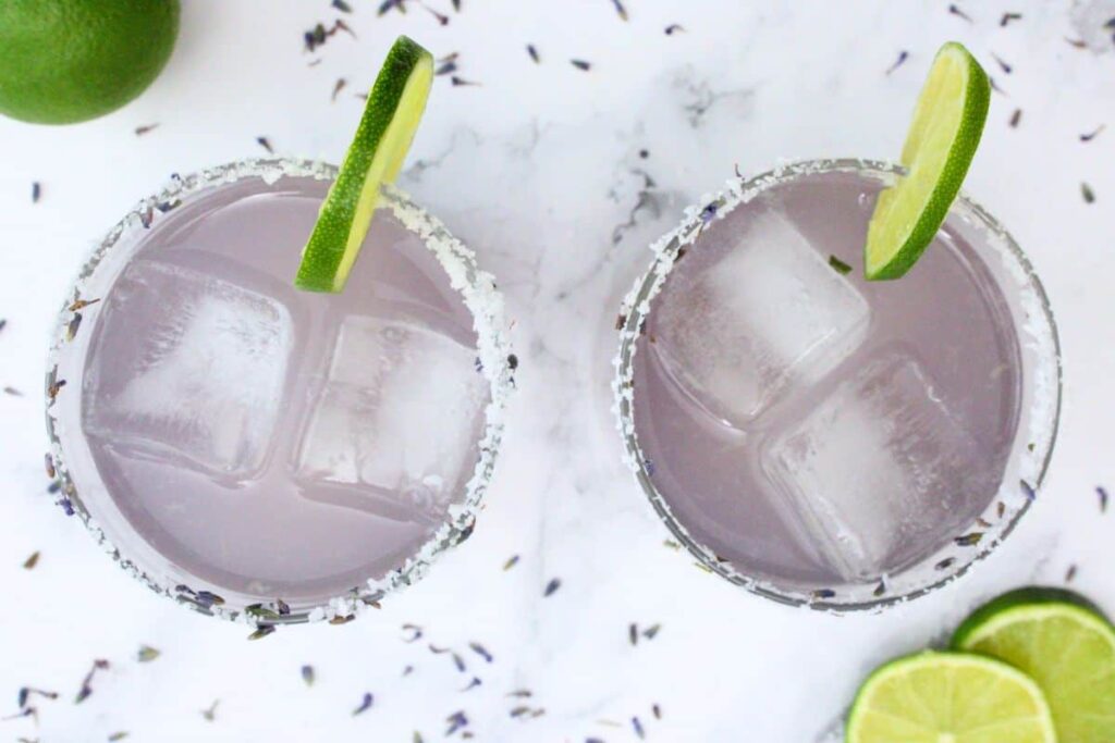 overhead view of two lavender margaritas with lime slice surrounded by lavender seeds and fresh lime