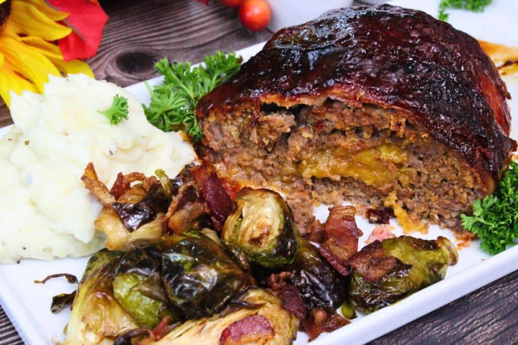 showing half of a bacon wrapped meatloaf stuff with cheese on a platter with mashed potatoes and bacon brussel sprouts