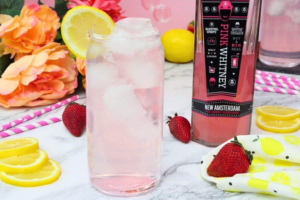 tall glass of pink whitney and sprite garnished with lemon next to pink whitney bottle