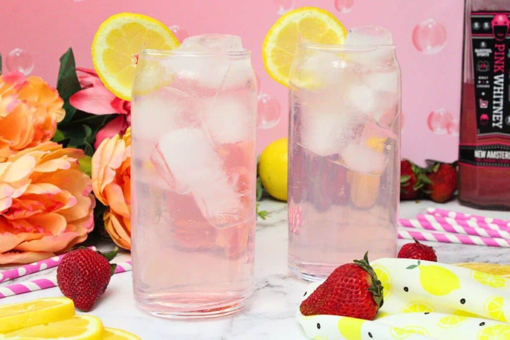 two pink whitney and sprite cocktails in tall glasses with a lemon slice in front of a pink background