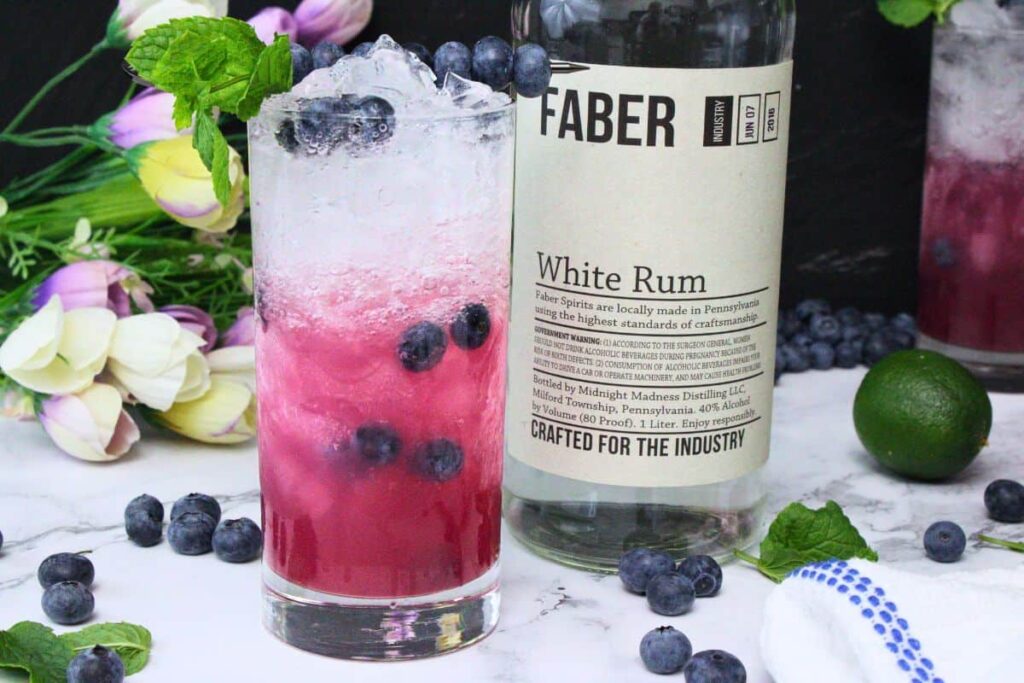 blueberry mojito in a tall glass garnished with mint next to rum bottle