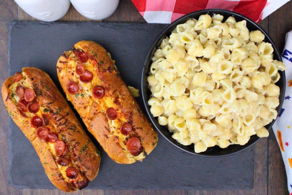 over head view of two pizza hot dogs with mini pepperoni next to bowl of mac and cheese on a slate board