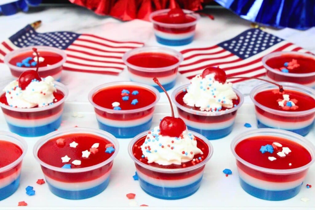 several red white and blue jello shots topped with whipped cream and a cherry surrounded by patriotic decor