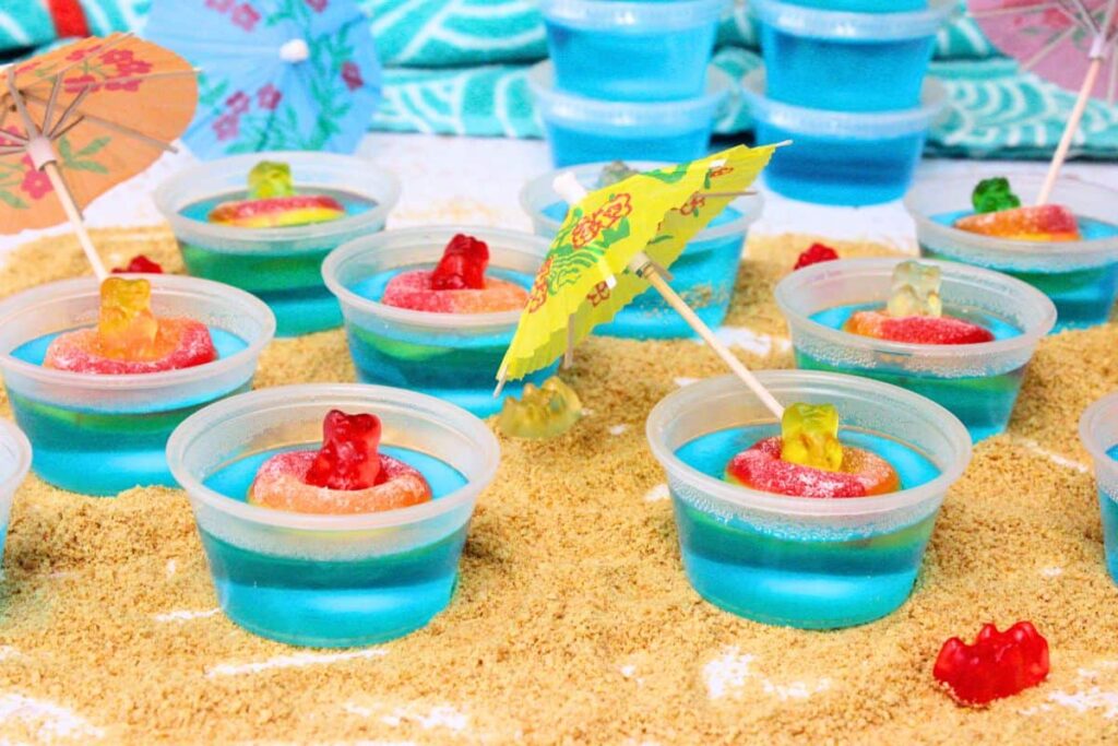 several pool party jello shots with gummy bears floating in peach rings on a graham cracker beach with cocktail umbrellas