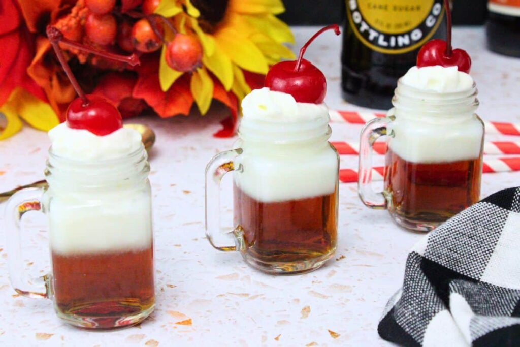 three root beer float shots in a line topped with whipped cream and a cherry