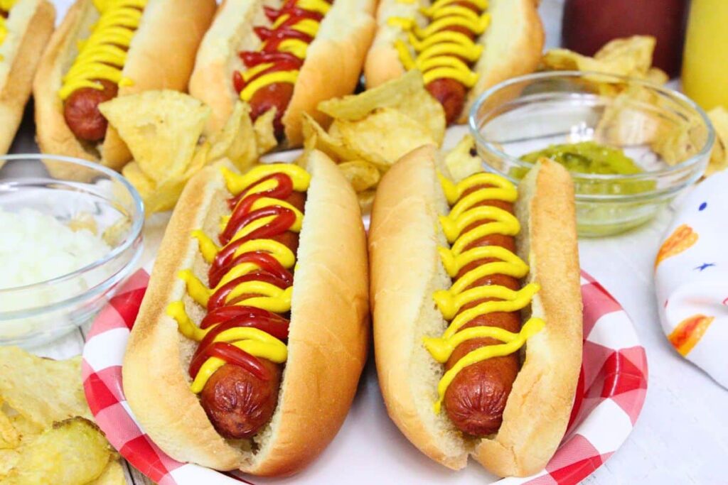 two air fryer hot dogs topped with ketchup and mustard on a summer plate surrounded by chips