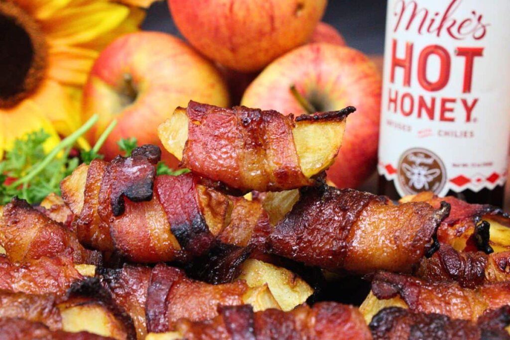 closeup view of air fryer bacon wrapped apples stacked up with a bottle of hot honey in the background
